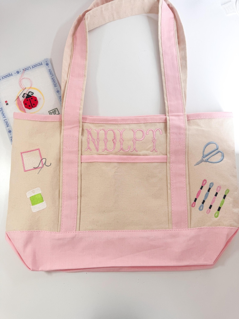 Op de afbeelding: Een beige canvas draagtas met roze accenten, met borduursels met naaigerei. De tas heeft roze handvatten en een roze bodem. De voorkant van de tas heeft de geborduurde letters "NOLPT". Extra geborduurde elementen zijn scharen, garenklosjes en een naald.