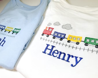 Könnte beinhalten: Zwei weiße T-Shirts mit gestickten Zugmotiven und Namen. Das Shirt links sagt "Noah" und das Shirt rechts sagt "Henry".