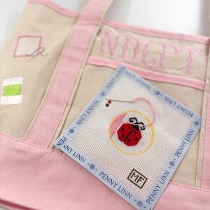 Op de afbeelding: Een beige draagtas met roze randen en handvatten. Een kleine vierkante naaldkunst met een lieveheersbeestjesontwerp is bevestigd. De tas heeft geborduurde tekst en een klein gestikt ontwerp. De naaldkunst heeft de tekst "PENNY LINN".