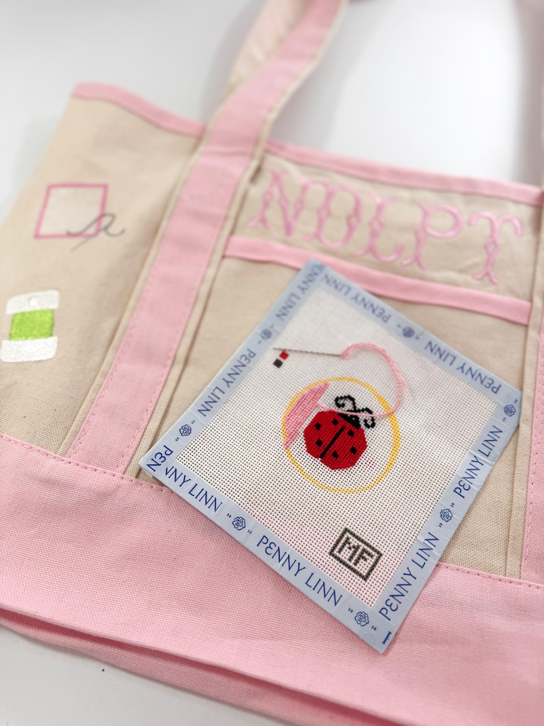Op de afbeelding: Een beige draagtas met roze randen en handvatten. Een kleine vierkante naaldkunst met een lieveheersbeestjesontwerp is bevestigd. De tas heeft geborduurde tekst en een klein gestikt ontwerp. De naaldkunst heeft de tekst "PENNY LINN".