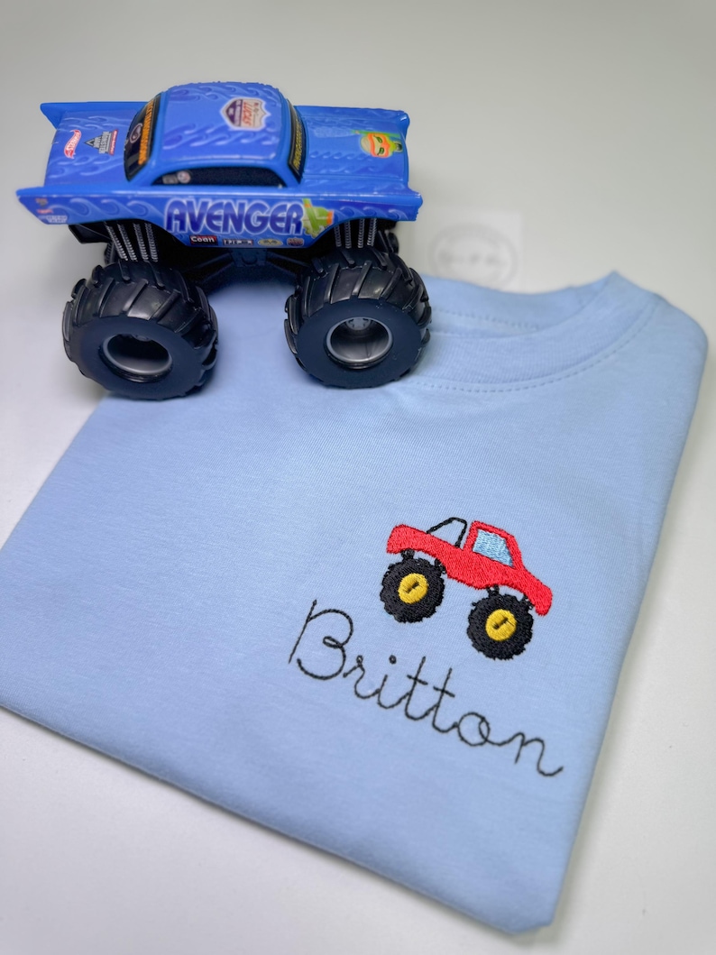 May include: A light blue t-shirt with a red monster truck embroidered on the front. The name "Britton" is embroidered below the truck.
