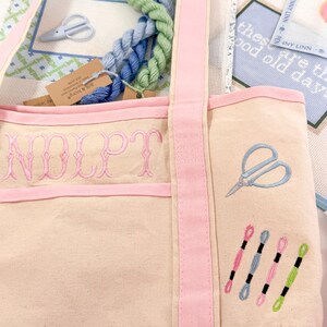 Op de afbeelding: Een beige canvas draagtas met roze randen en bandjes, geborduurd met de letters "NOLPT" in roze. De tas heeft een blauw schaarontwerp en verschillende spoelen garen in roze, blauw en groen. Binnenin zitten strengen blauw en groen garen.