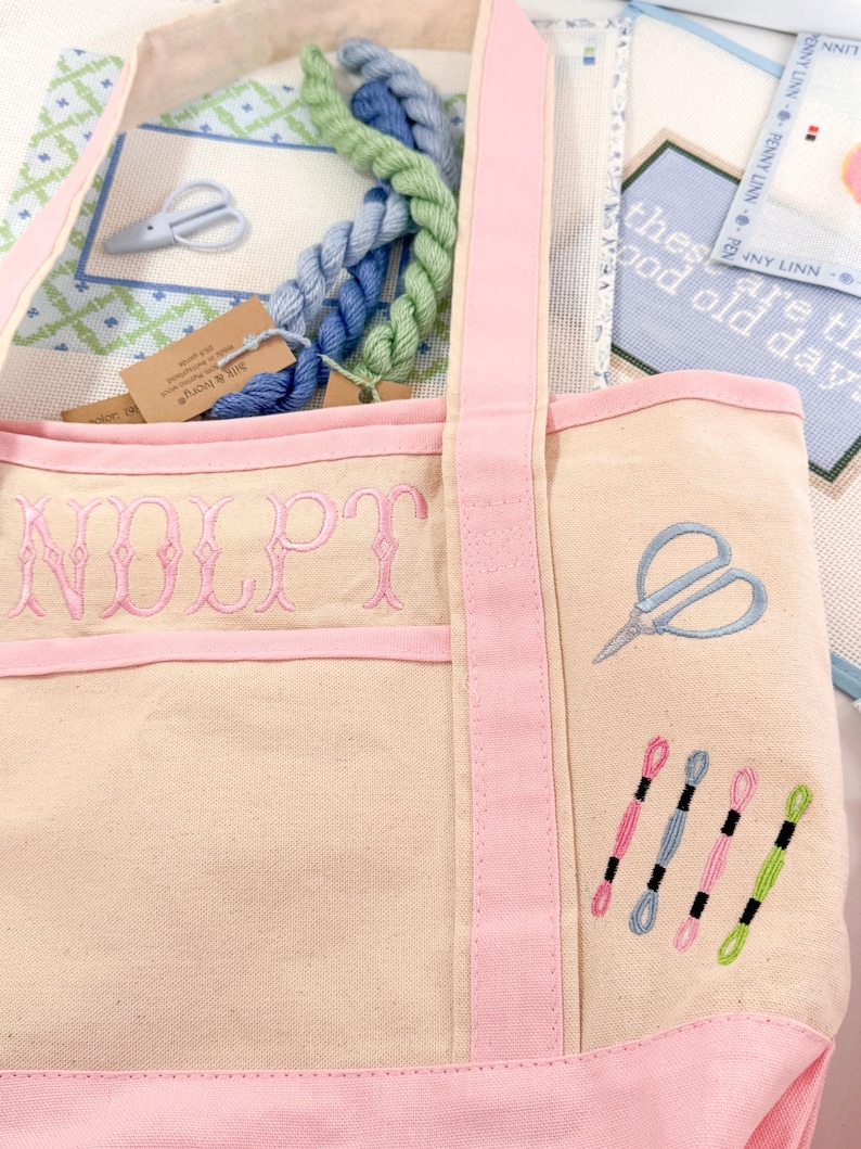 Op de afbeelding: Een beige canvas draagtas met roze randen en bandjes, geborduurd met de letters "NOLPT" in roze. De tas heeft een blauw schaarontwerp en verschillende spoelen garen in roze, blauw en groen. Binnenin zitten strengen blauw en groen garen.