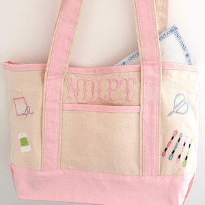 Op de afbeelding: Beige draagtas met roze accenten en borduursels met een naaithema. De tas heeft roze handvatten en een roze bodem. De initialen "NOLPT" zijn op de voorkant geborduurd. Een borduurcanvas is zichtbaar in de zak.