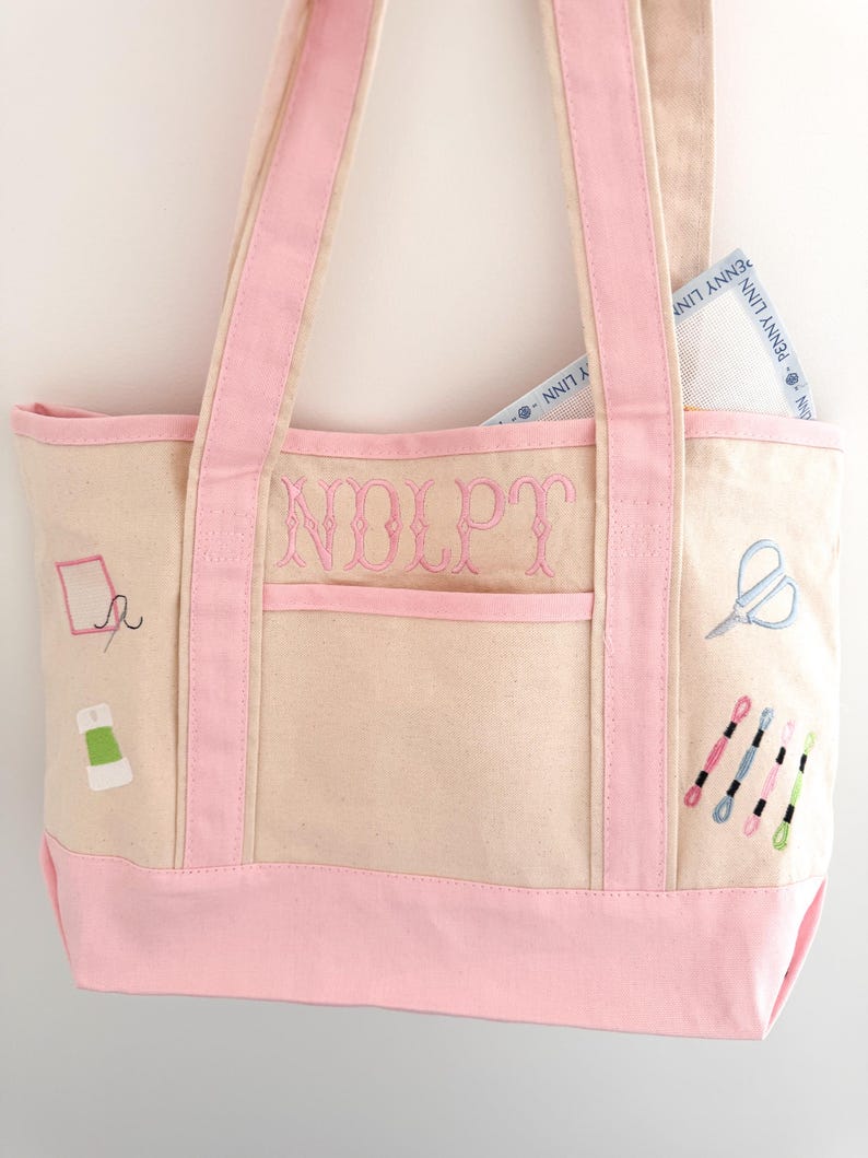 Op de afbeelding: Beige draagtas met roze accenten en borduursels met een naaithema. De tas heeft roze handvatten en een roze bodem. De initialen "NOLPT" zijn op de voorkant geborduurd. Een borduurcanvas is zichtbaar in de zak.