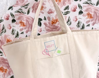 May include: A cream-colored canvas tote bag featuring two sturdy handles and an exterior pocket. The front is embroidered with a light blue square frame containing the text "NDLPT" in pink, alongside a pink pair of scissors and a green spool.