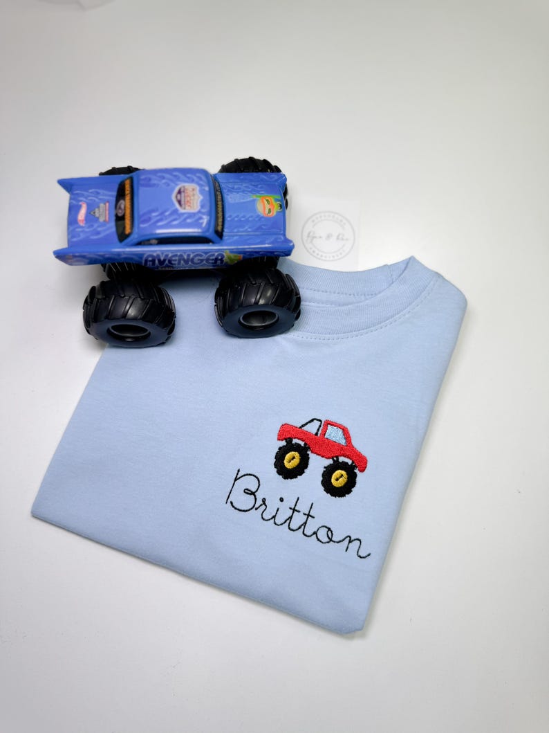 May include: Light blue t-shirt with an embroidered red monster truck and the name Britton.
