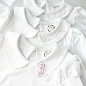 Puede incluir: Cuatro camisas blancas con cuello y bordados rosas. Las camisas están personalizadas con los nombres Olivia, Kennedy y un monograma "B".