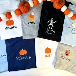 Puede incluir: Una colección de ocho camisetas en blanco, gris, negro y azul claro, cada una con diseños bordados de Halloween que incluyen calabazas, un caramelo de maíz, un esqueleto, un fantasma y un cubo de calabaza, con varios nombres y frases.