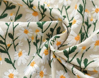 May include: A beige fabric featuring an embroidered pattern of white daisy flowers with yellow centers and green stems and leaves. The textile is gathered in folds, showcasing the detailed stitching of the floral design across the surface.