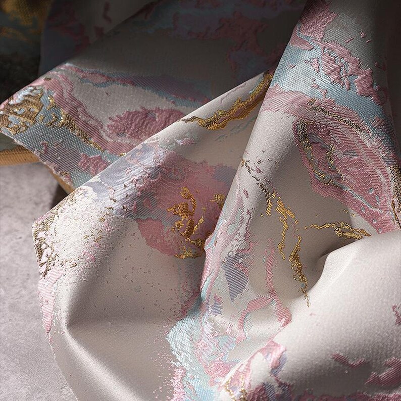 May include: Close-up of a soft, white fabric with a pink, blue, and gold marbled pattern. The fabric has a subtle sheen and a textured surface.