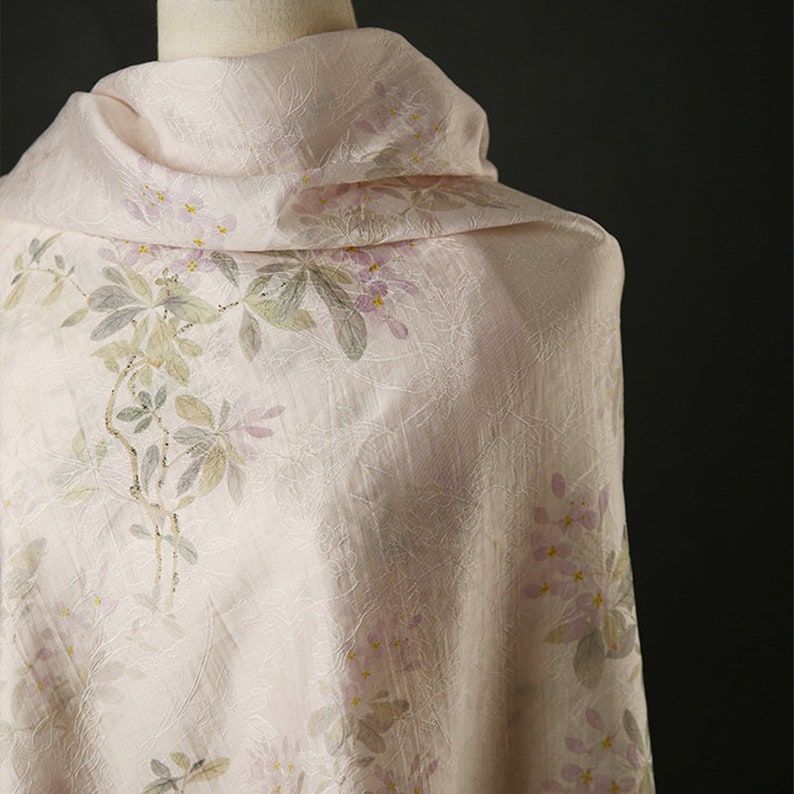Pu&ograve; includere: Un tessuto rosa pallido con un delicato motivo floreale. I fiori sono di un viola chiaro e le foglie sono di un verde tenue. Il tessuto ha una texture sottile.