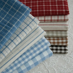 May include: A stack of folded fabric squares in various patterns and colors. The fabrics include red, blue, white, brown, and black checkered and striped designs. The fabrics are stacked on a white lace surface.
