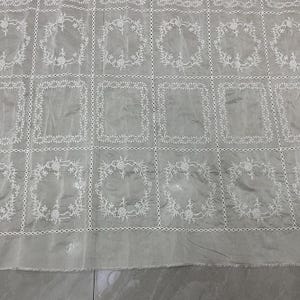 May include: A light gray fabric featuring a grid pattern formed by white embroidered lines. Each square in the grid contains a white embroidered floral wreath design. The fabric has a semi-sheer quality with a visible texture and a raw bottom edge.