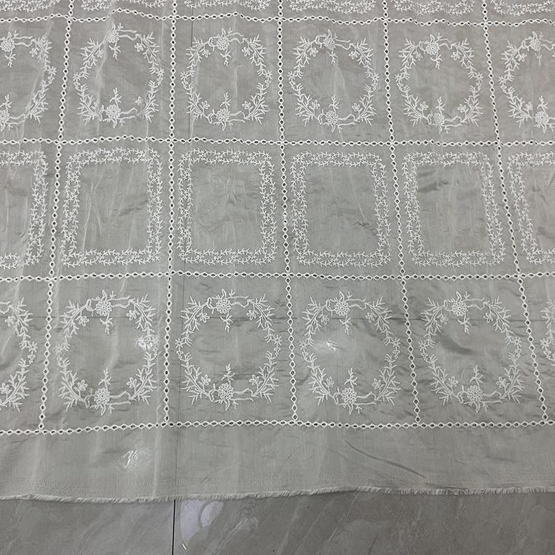 May include: A light gray fabric featuring a grid pattern formed by white embroidered lines. Each square in the grid contains a white embroidered floral wreath design. The fabric has a semi-sheer quality with a visible texture and a raw bottom edge.
