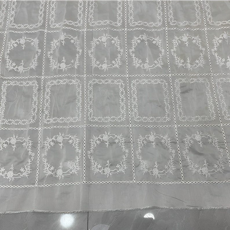 May include: A sheer light gray fabric featuring a repeating grid pattern of white embroidered rectangles and circular floral wreaths. The embroidery is delicate and creates a structured geometric design across the translucent textile surface.