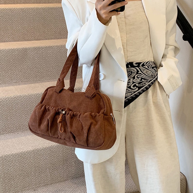 May include: A brown corduroy handbag with a drawstring closure and a silver accent. The bag has a structured shape and a top handle.