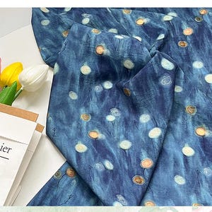 May include: A folded piece of fabric with a blue watercolor-style background and a pattern of white and gold circular shapes. The fabric is draped over a white surface. A paper bag with the word "papier" is visible.