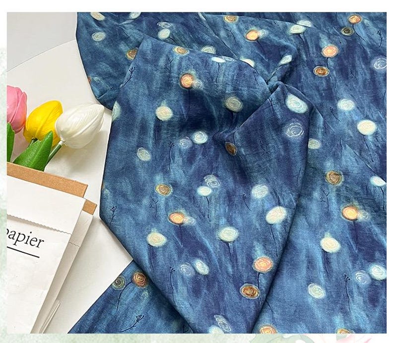 May include: A folded piece of fabric with a blue watercolor-style background and a pattern of white and gold circular shapes. The fabric is draped over a white surface. A paper bag with the word "papier" is visible.