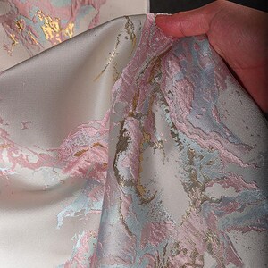 May include: Close-up of a white fabric with a pink, blue, and gold marble pattern. The fabric has a textured, raised surface.