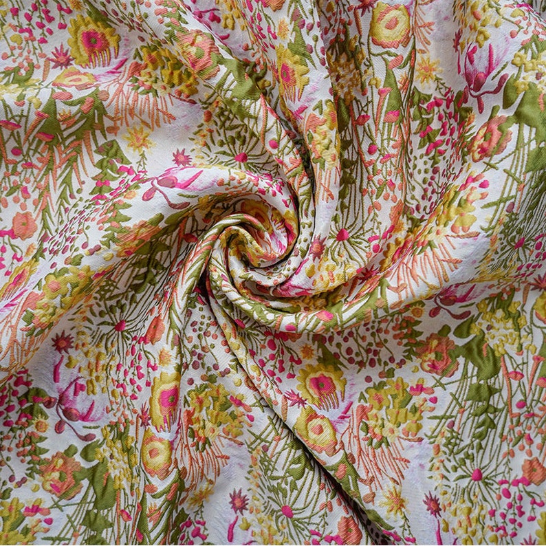 May include: A white fabric with a floral pattern in shades of pink, yellow, and green. The fabric is textured and has a slightly raised design.