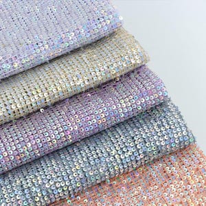 Può includere: Primo piano di diversi campioni di tessuto ricoperti di paillettes iridescenti. I tessuti sono impilati, rivelando strati di trame scintillanti nelle tonalità lavanda, argento e pesca. Le paillettes sono cucite su un supporto a rete.