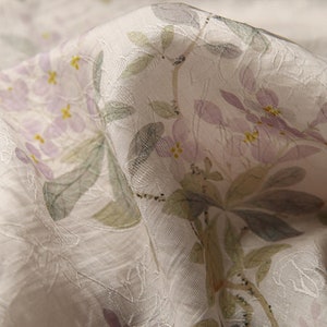 Pu&ograve; includere: Primo piano di un tessuto bianco con un motivo floreale. I fiori sono di colore viola chiaro con centri gialli e sono circondati da foglie verdi. Il tessuto ha una superficie testurizzata.
