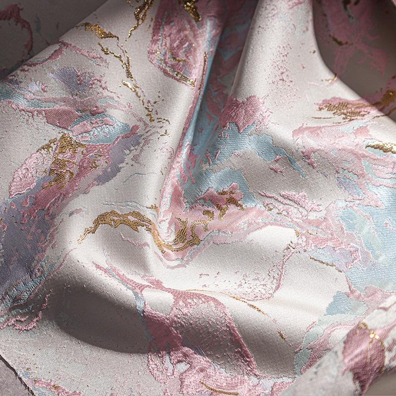 May include: A close-up of a fabric with a pink, blue, and gold marble pattern. The fabric is soft and has a subtle sheen.