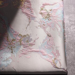 May include: Close-up of a white fabric with a pink, blue, and gold marbled pattern. The fabric has a textured surface.
