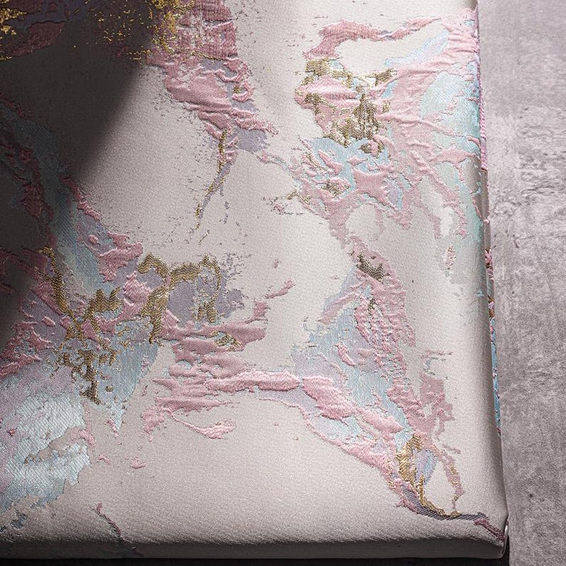 May include: Close-up of a white fabric with a pink, blue, and gold marbled pattern. The fabric has a textured surface.