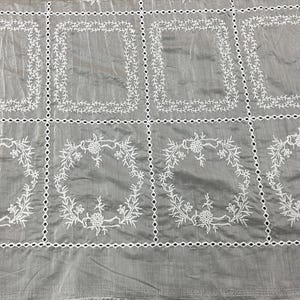 May include: A grey fabric featuring a grid pattern created by white embroidered eyelet borders. The top row displays rectangular frames of white floral vines, while the bottom row features circular wreaths of white flowers and leaves.