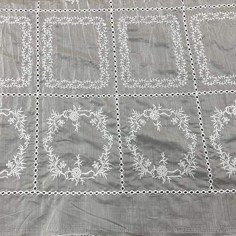 May include: A grey fabric featuring a grid pattern created by white embroidered eyelet borders. The top row displays rectangular frames of white floral vines, while the bottom row features circular wreaths of white flowers and leaves.