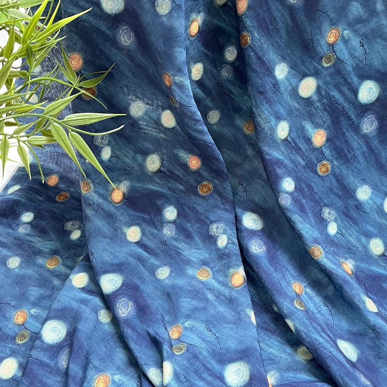May include: A flowing fabric with a deep blue background, adorned with a pattern of white and orange-toned circular designs. The fabric has a watercolor-like effect, with subtle variations in color and texture. The design includes small, delicate black lines.