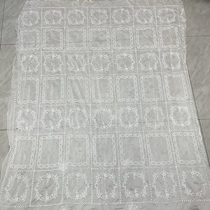 May include: A white rectangular lace tablecloth featuring a repeating grid pattern of embroidered floral wreaths and solid rectangular frames. The fabric is sheer and lightweight, with a delicate stitched border separating each individual square section.