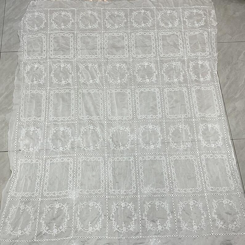 May include: A white rectangular lace tablecloth featuring a repeating grid pattern of embroidered floral wreaths and solid rectangular frames. The fabric is sheer and lightweight, with a delicate stitched border separating each individual square section.