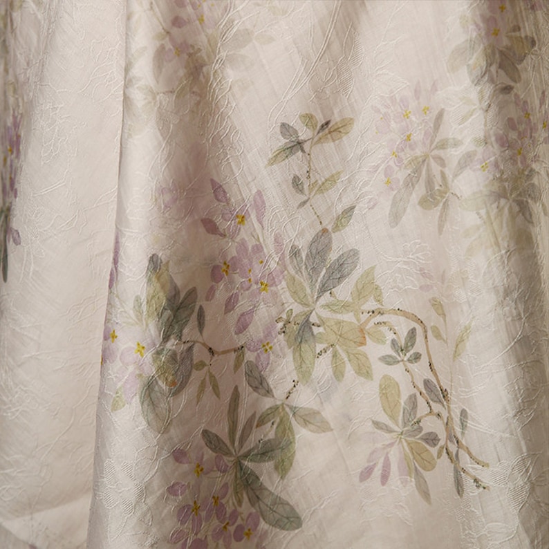 Pu&ograve; includere: Un tessuto bianco con un delicato motivo floreale. I fiori sono di un rosa pallido e le foglie di un verde chiaro. Il tessuto ha una texture sottile.