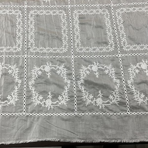 May include: A grey fabric featuring a grid of white embroidered eyelet borders. The top row displays rectangular frames with vine patterns, while the bottom row features circular floral wreaths. The fabric has a visible weave and a raw bottom edge.
