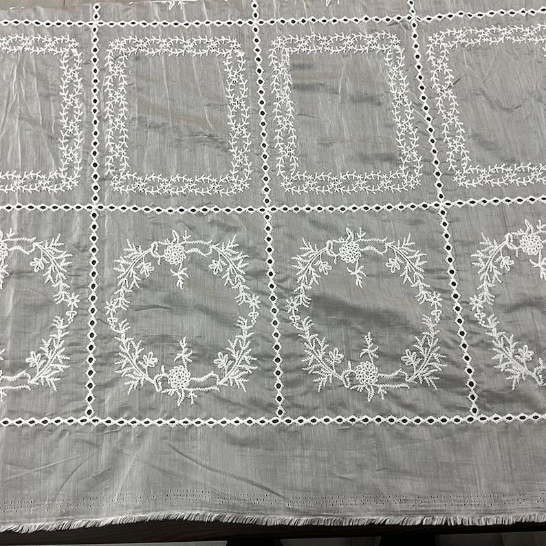 May include: A grey fabric featuring a grid of white embroidered eyelet borders. The top row displays rectangular frames with vine patterns, while the bottom row features circular floral wreaths. The fabric has a visible weave and a raw bottom edge.