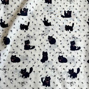 Pode incluir: Tecido branco com um padrão repetido de ilustrações de gatos pretos. Cada gato tem um laço rosa. Pequenos detalhes florais e laços pretos estão espalhados pelo fundo. Os gatos estão em várias poses, criando um design lúdico e caprichoso.