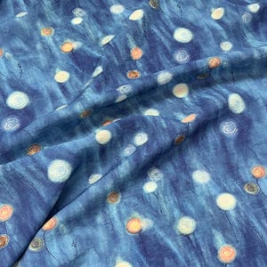 May include: A close-up of a blue fabric with a pattern of white and orange floral designs. The fabric has a textured appearance with vertical brushstrokes, creating a watercolor effect. The floral elements are scattered across the surface.