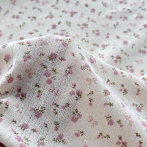 May include: Close-up of white fabric with a delicate floral pattern. Small pink roses and bows are scattered across the material. The fabric has a textured, open weave, suggesting a lightweight textile.