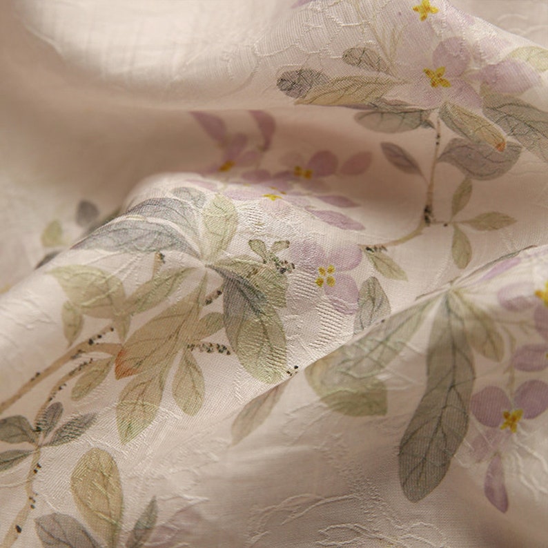 Pu&ograve; includere: Primo piano di un tessuto morbido e bianco con un delicato motivo floreale. I fiori sono di un viola pallido con centri gialli e sono sparsi su tutto il tessuto. Il tessuto ha una texture sottile, che gli conferisce un aspetto leggermente rialzato.