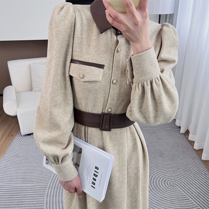 May include: A beige corduroy dress with a brown leather collar and belt. The dress has a button-up front and long sleeves with puffed shoulders. The dress has a large pocket on the left side with a brown leather trim.