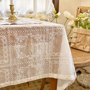 May include: A white rectangular tablecloth featuring a grid pattern with intricate floral embroidery and cutwork details. The fabric has a sheer, textured appearance with repeating circular botanical motifs within each square section.