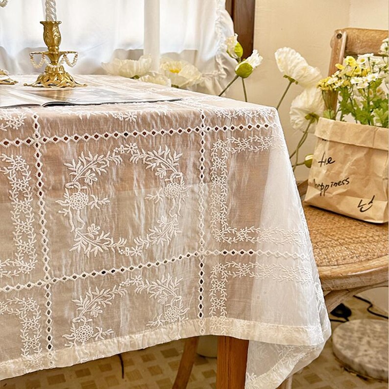 May include: A white rectangular tablecloth featuring a grid pattern with intricate floral embroidery and cutwork details. The fabric has a sheer, textured appearance with repeating circular botanical motifs within each square section.