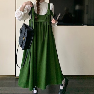 May include: A green corduroy overall dress with thin straps and a bow detail at the shoulders. The dress has a loose fit and falls to the mid-calf. The model is wearing black shoes with white accents.