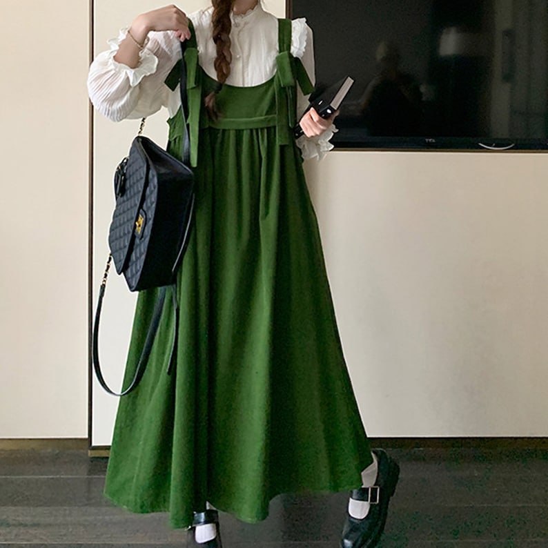 May include: A green corduroy overall dress with thin straps and a bow detail at the shoulders. The dress has a loose fit and falls to the mid-calf. The model is wearing black shoes with white accents.