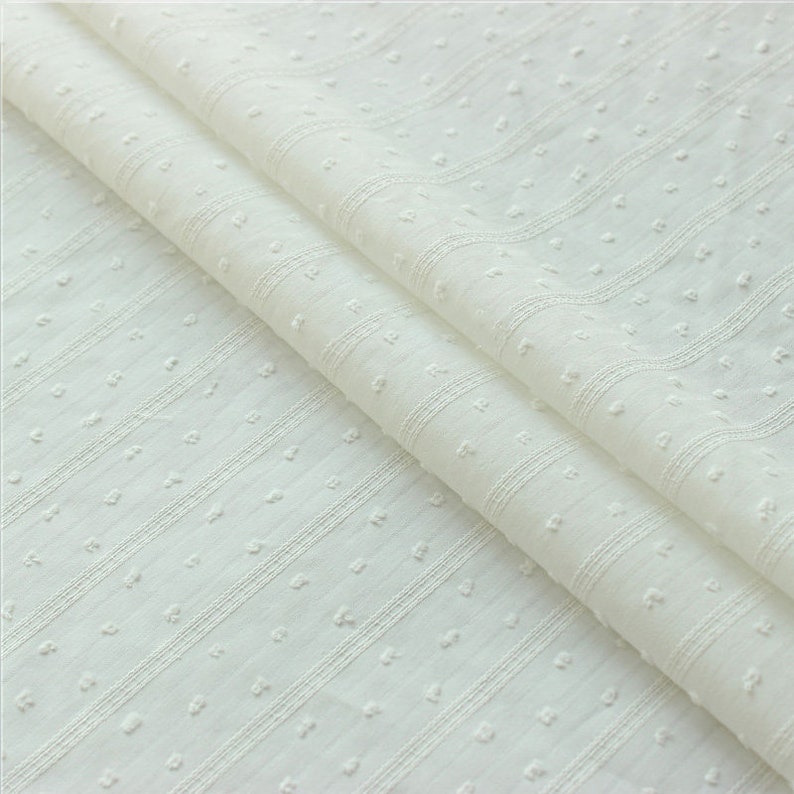 May include: White cotton fabric with a textured, dotted pattern and thin, horizontal stripes. The fabric is folded in half, showing the pattern on both sides.