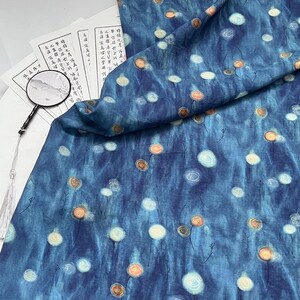 May include: A piece of blue fabric with a watercolor-like design featuring white and orange floral patterns. The fabric is draped over a stack of papers with Asian calligraphy and a magnifying glass.