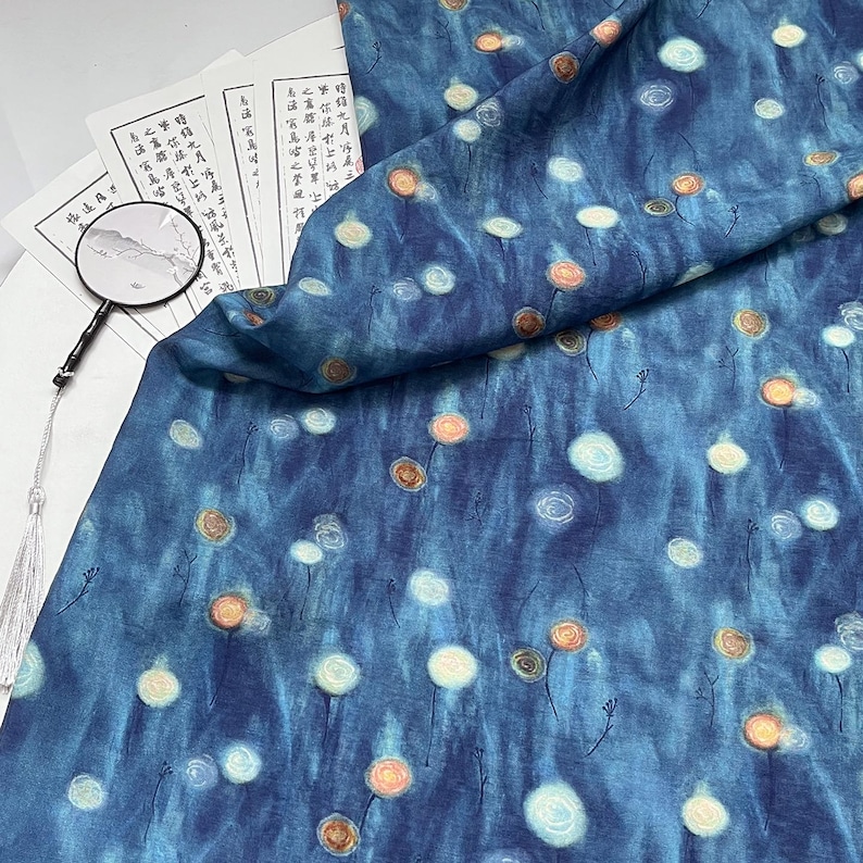 May include: A piece of blue fabric with a watercolor-like design featuring white and orange floral patterns. The fabric is draped over a stack of papers with Asian calligraphy and a magnifying glass.